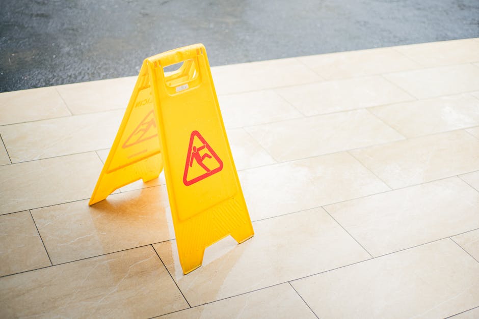 A yellow caution wet floor sign stands on beige ceramic tile flooring inside a room, indicating a recent cleaning or potential hazard. The sign displays a red pictogram of a person slipping. The polis