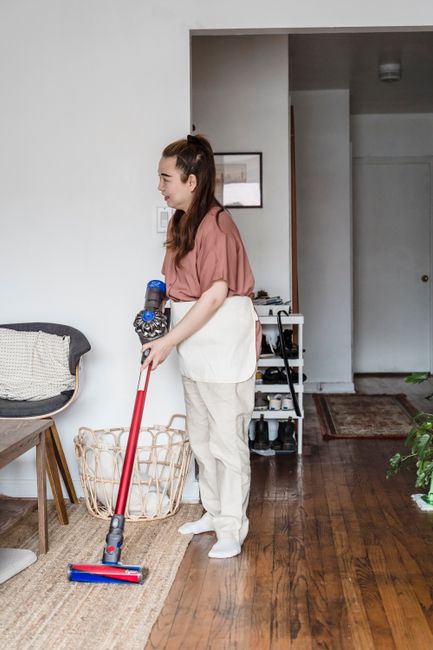 A woman with long brown hair tied back, wearing a pink short-sleeved top and beige trousers with an apron, is using a cordless vacuum cleaner with a red handle and a blue cleaning head to clean a wood
